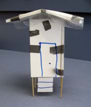 Photo shows a small house made of foam core board on toothpick stilts, with a low-peaked plastic-covered roof with wide overhangs.