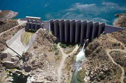 Aerial photo shows a concrete structure blocking a river flow and holding back a supply of water. From above, the structure shape looks like a series of scallops across the river width.