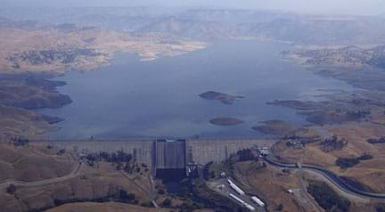 Aerial photo shows a wide and straight concrete structure blocking a river flow and holding back a supply of water. From above, the structure shape looks like a wall across the river width, tipped towards the resulting reservoir.