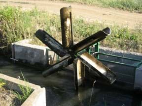 Photo shows a metal waterwheel spinning in a ditch stream as moving water catches in its six curved blades.