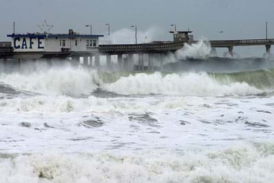 Violent and frothy waves splash around the walkway and columns of an ocean-side pier, as well as the shallow water near the shore.