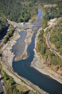 Aerial photo shows a narrow stream of water flowing unobstructed through one side of a river in which the concrete dam is gone while a bulldozer moves piles of dirt to block the water's passage through the remaining half of the dam.