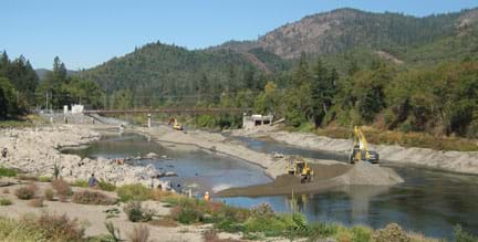 Landscape photo shows a pile of soil positioned lengthwise a few hundred meters down the middle of a river above a dam. Half the river flows over a concrete embankment and half flows free. A bulldozer pushes dirt into the river to block the water's route towards the remaining half dam, directing all the water to the free-flowing side.