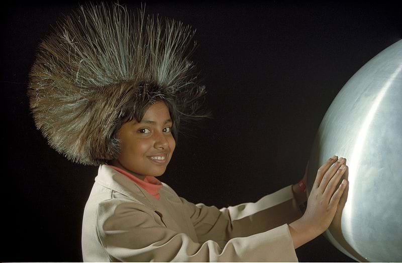 This photograph shows the surprise in a young person's face and their long hair standing on end around their head as they touch a large, shiny silver orb in front of them.