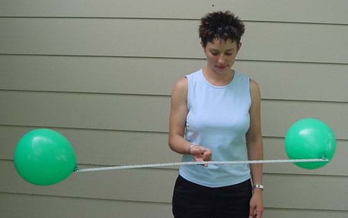 Photograph of a woman holding a meter stick, with inflated balloons attached on each end, perfectly balanced on one finger.