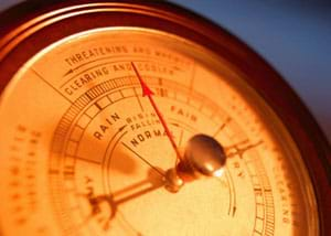 A close-up photograph shows the face of a barometer instrument with an arrow hovering between the markings for Rain and Fair.