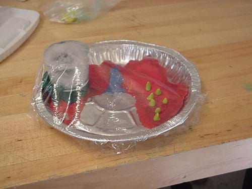 A photograph shows an aluminum pie tin with clay shapes in the bottom, a chunk of ice sitting on part of the clay, and the entire tin enclosed with plastic wrap.
