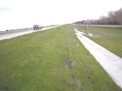 Photo shows a grassy area to the side of a highway, sloping down to a concrete collection drain. 