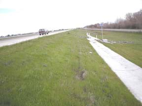 Photo shows a grassy area to the side of a highway, sloping down to a concrete collection drain.