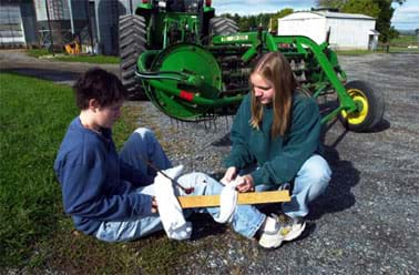 One person uses rags to tie a piece of wood to another person's leg to immobilize it.