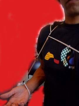A photo shows a person's torso and right arm, with a thin rope around his wrist, connected by a rubber band to another rope that runs around his shoulder and across his chest.