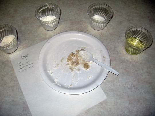 A photo shows amounts of flour, sugar, cornstarch and oil in clear plastic cups arranged around a paper plate and plastic spoon on a desktop.