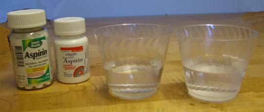 Photo shows two bottles, of regular and enteric-coated aspirin, next to clear cups, each half filled with clear liquid.
