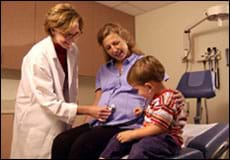 A pregnant woman and a child in an exam room, with a doctor pressing a stethoscope to the woman's abdomen as the child listens with the stethoscope.