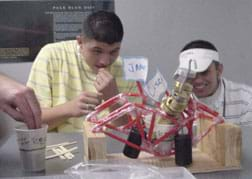 A photo shows two students intently watching a collapsing model bridge.