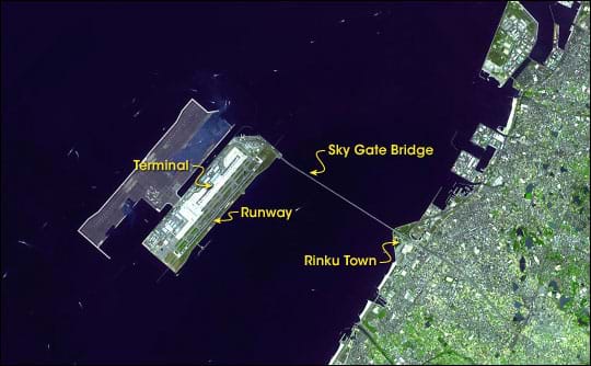 Aerial satellite image shows urban shoreline, blue bay waters, and long bridge connecting shore to a rectangular island containing airport terminal and runways.