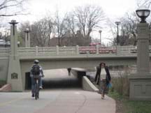 Photo shows bikers and walkers going under a bridge and cars traveling over a bridge.