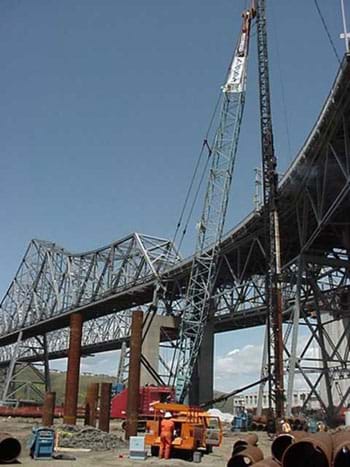 Photo shows a crane lifting a steel truss from the ground to a bridge deck.