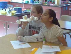 Photo of one girl holding a paper plate and paper cup contraption, and another girl blowing on it. 