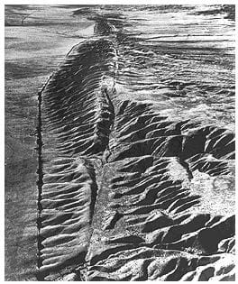 A black and white aerial photograph shows what looks like a slice through a ridge of desert mountains and foothils. It is the San Andreas fault in the Carrizo Plan in central California.