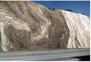 A photograph shows a rock cliff with curved and folded layers of sedimentary rocks, as evidenced by the colored layers of the formation.