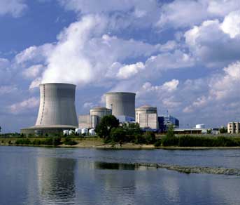 A photograph of a nuclear power plant. Shown is an industrial-looking plant with two large concrete stacks with plumes of steam escaping. The plant sits on the shore of a body of water, and many buildings surround the two stacks.