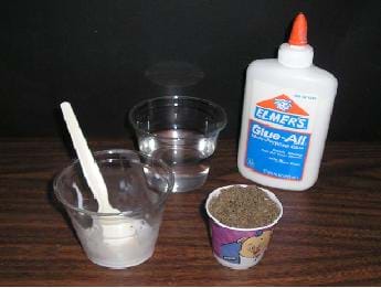 Photo shows a counter top with a bottle of glue, a Dixie cup filled with brown material, a cup of water and a cup with white glue and a spoon.