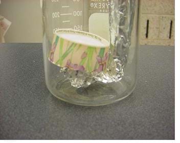 A photograph shows the bottom of a glass jar containing a rolled aluminum foil column bent at a 90-degree angle across the bottom of the jar. Past the bend, some of the foil is crumpled against the bottom of the glass jar. On top of the crumpled aluminum foil is an upside down paper cup bottom.
