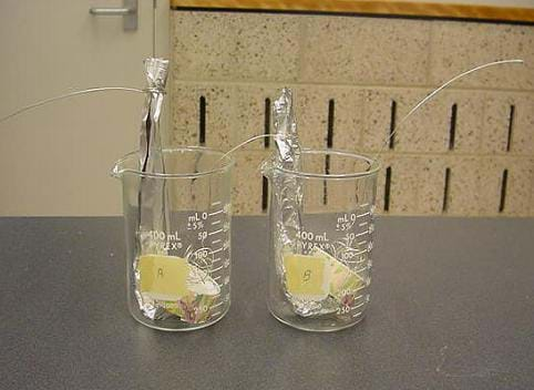 A photograph of the activity set up. On the left, a glass container marked "A" holds a rolled aluminum foil column with a 90-degree angle and the part of the foil after the angle crumpled against the bottom of the glass jar. On top of the crumpled piece of aluminum is an upside down bottom of a small paper cup. A coiled piece of copper wire sits on top of the paper cup bottom. The other side of the copper wire is connected to the top of the aluminum foil column that is situated in a second glass container, labeled "B," located to the right of container "A." Container B has the same setup described for container A, however, the coiled copper wire on top of the paper cup leads out of the container and is not connected to anything.