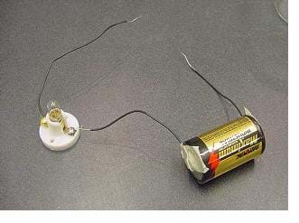 A photograph of the activity's conductivity tester for solids. To the positive terminal of a D-cell battery, a short piece of wire is connected with masking tape. The other end of the wire is connected to the terminal of a light bulb holder. A second wire is connected to the negative terminal of the D-cell battery with masking tape and the other end of this wire is not connected to anything. A third wire is connected to the opposite and unused terminal of the light bulb holder and the other end of this wire is not connected to anything.