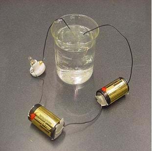 A photograph displays a conductivity tester for liquids and solutions. Two D-cell batteries are connected in series. A short wire and masking tape connects the positive terminal of one battery to the negative terminal of a second battery. The positive terminal of the second battery is connected to a light bulb holder with a second short wire. A third wire is connected to the opposite, unused terminal of the light bulb holder and the other end of the third wire is submersed in a solution within a beaker. A fourth wire is connected to the negative terminal of the first battery and the other end of the wire is also submersed in the solution in the beaker.