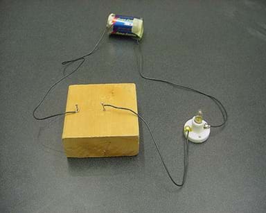 Photo shows the basic setup of a simple switch. One wire connects the positive terminal of the D-cell battery to one of the light bulb holder terminals. A second wire connects the negative terminal of the D-cell battery to a nail hammered into a block of wood. A third wire connects the second light bulb holder terminal to a second nail hammered into the wood, 2 cm from the first nail.