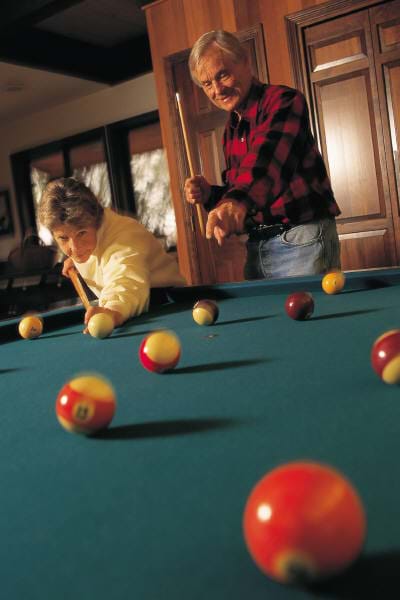 A photograph of a man and woman playing billiards, showing a scattering of balls across the pool table playing surface.