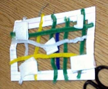 A picture of an example air filter to be used in the filter testing apparatus. Shown is an index card with a big square cut in the middle and pipe cleaners taped in a criss-cross pattern across the hole.
