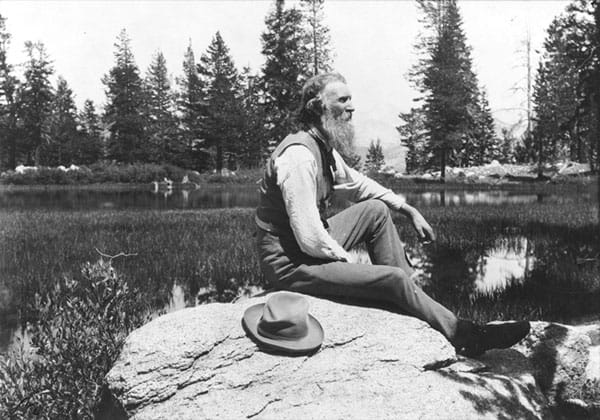 A portrait of John Muir, facing right, seated on a rock with lake and trees in background.