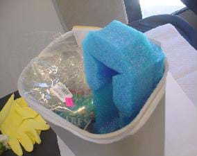 Photo shows a small plastic trash can filled with plastic wrap, a soda can and packing foam. Yellow rubber gloves lay nearby.