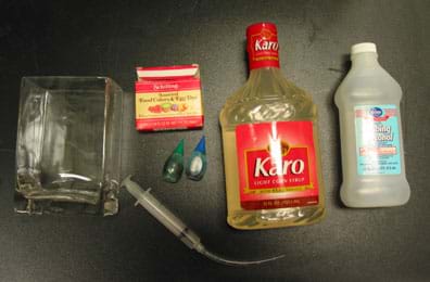 Bottles of corn syrup and alcohol, syringe with tube connected, box of liquid food coloring in small plastic squeeze bottles, square glass container.