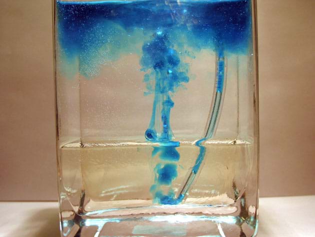 Photo of corn syrup on bottom of container and water on top with the syringe and tube inserted into the bottom of the container with blue dye rising up from end of tubing.