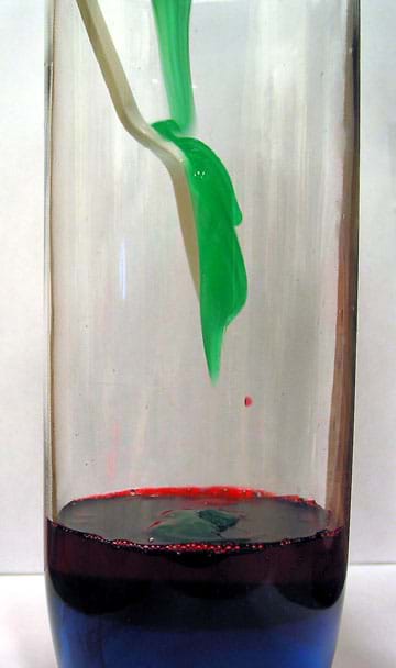 Photo shows a stream of green liquid hitting the back of a spoon midway inside a tall drinking glass as it is being poured onto a layer of red and blue fluids already in the container.