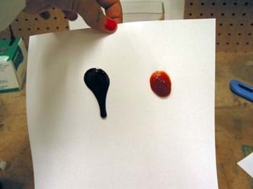  Photo shows a hand holding a piece of paper upright by the edge. On the paper, a dark drop slides further down the paper than a red drop.