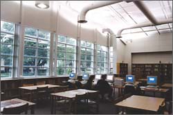 In a classroom with tables, chairs, desks and computers, light comes in through large windows and a round ceiling opening.