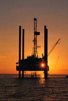 Photo shows silhouette of poles and a platform holding a drilling rig above the surface of the ocean at sunset.