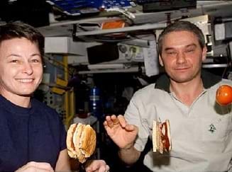 Photo shows two astronauts eating floating sandwiches and fruit aboard the International Space Station (ISS).