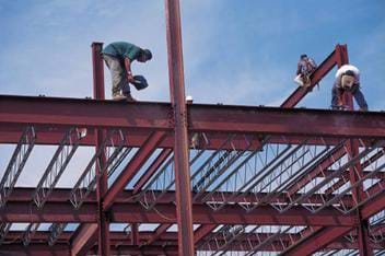 A photograph of I-beams being installed for a structure that is under construction. Three construction workers are standing on the beams working.