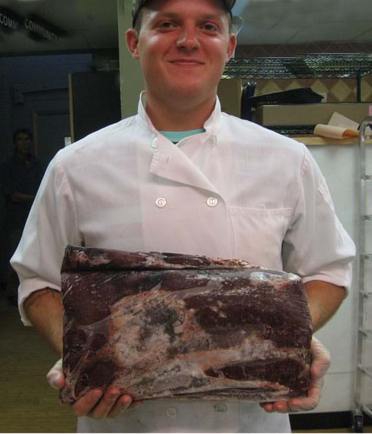 Photo shows a man in a white button jacket holding a reddish-brown slab of meat that is the size of a small briefcase.