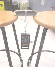 This photograph shows a seven-inch clay I-beam sitting on the edges of two stool seats, spanning a six-inch gap between the two stools, with a weight hanging from a loop of string placed around the middle of the beam.