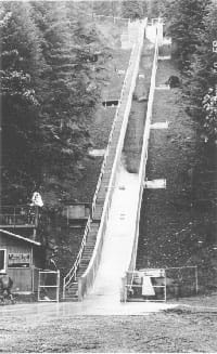 Black/white photo of a concrete chute up a hillside with a rush of soil coming down, banked by a set of stairs to the top.