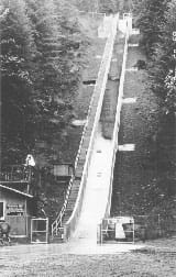 Black/white photo of a concrete chute up a hillside with a rush of soil coming down, banked by a set of stairs to the top.