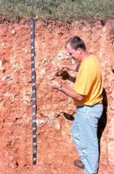Photo shows a man standing next to a cut-away section of soil as tall as he is, with an attached 76-inch (193-cm) measuring tape, and a short amount of grass and top soil above a deep, reddish-colored bedrock layer.