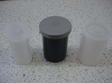 A photograph shows three types of film canisters sitting on a counter top: (left) a translucent plastic container and lid that are elliptical in shape; (center) a 35 mm photographic film canister made of black plastic with a gray overlapping cap; (right) a 35 mm photographic film canister made of translucent plastic with an inset fitted cap.